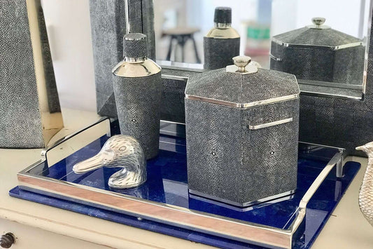 A chic cocktail shaker with a charcoal grey shagreen exterior and stainless steel interior, placed on a blue surface alongside other matching bar accessories.